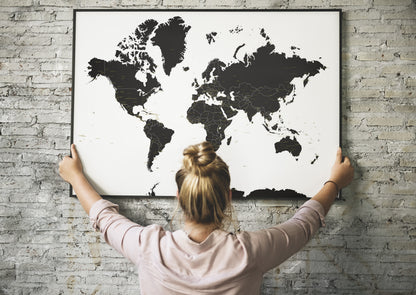 A woman with her hair in a bun hangs a large black-and-white world map poster on a light gray brick wall, viewed from behind her.