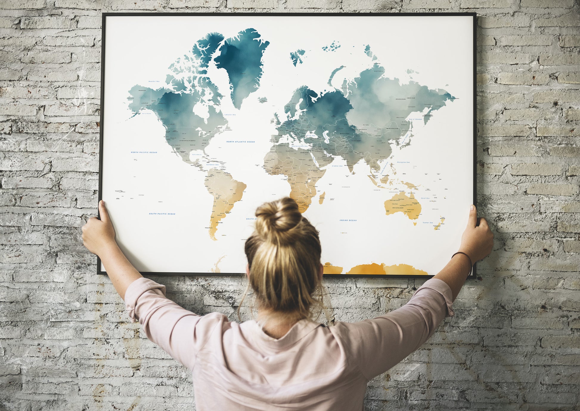 A woman with her hair in a bun hangs a large framed world map on a gray brick wall, viewed from behind. The map features a watercolor design with blue oceans and gold landmasses.
