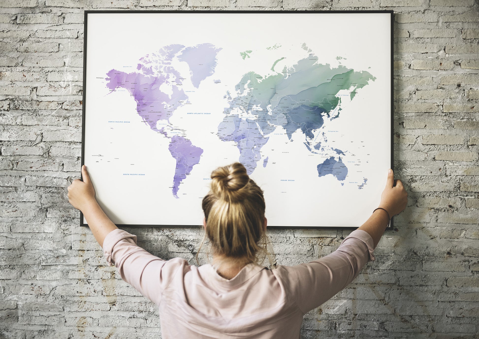 A woman with her hair in a bun hangs a large framed world map poster on a light gray brick wall, viewed from behind her.