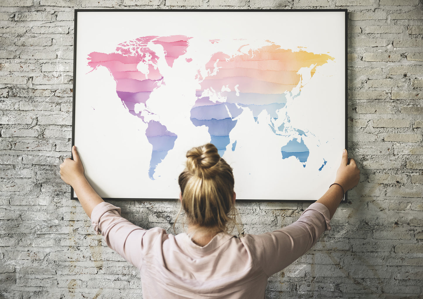 A woman with her hair in a bun hangs a large framed world map poster on a gray brick wall. The map features a colorful geometric, layered design with pink, purple, blue, and orange tones on a white background.