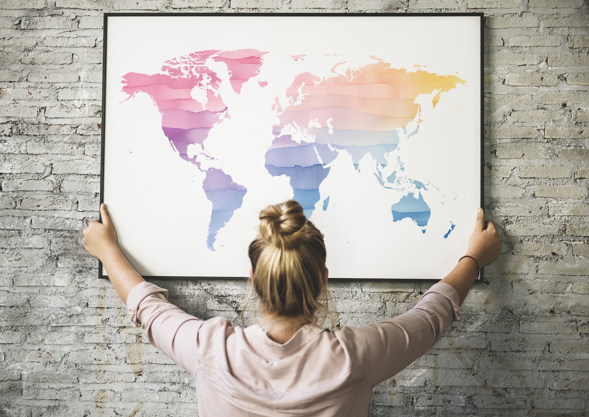 A woman with her hair in a bun hangs a large framed world map poster on a gray brick wall. The map features a colorful geometric, layered design with pink, purple, blue, and orange tones on a white background.