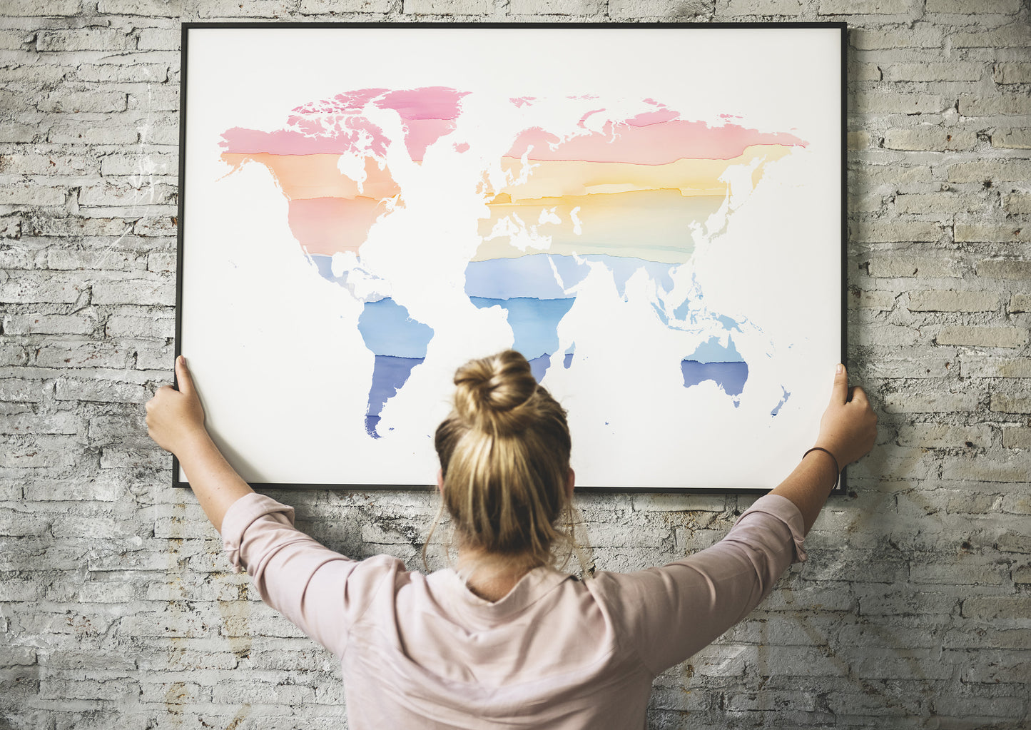 A woman with her hair in a bun hangs a large framed world map poster on a gray brick wall. The map features watercolor-style continents in pink, orange, yellow, and blue on a white background.