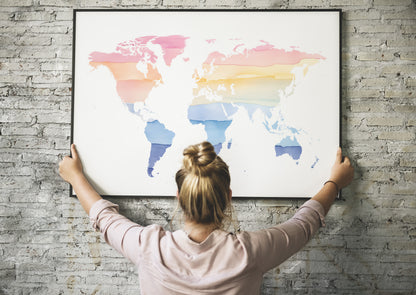 A woman with her hair in a bun hangs a large framed world map poster on a gray brick wall. The map features watercolor-style continents in pink, orange, yellow, and blue on a white background.
