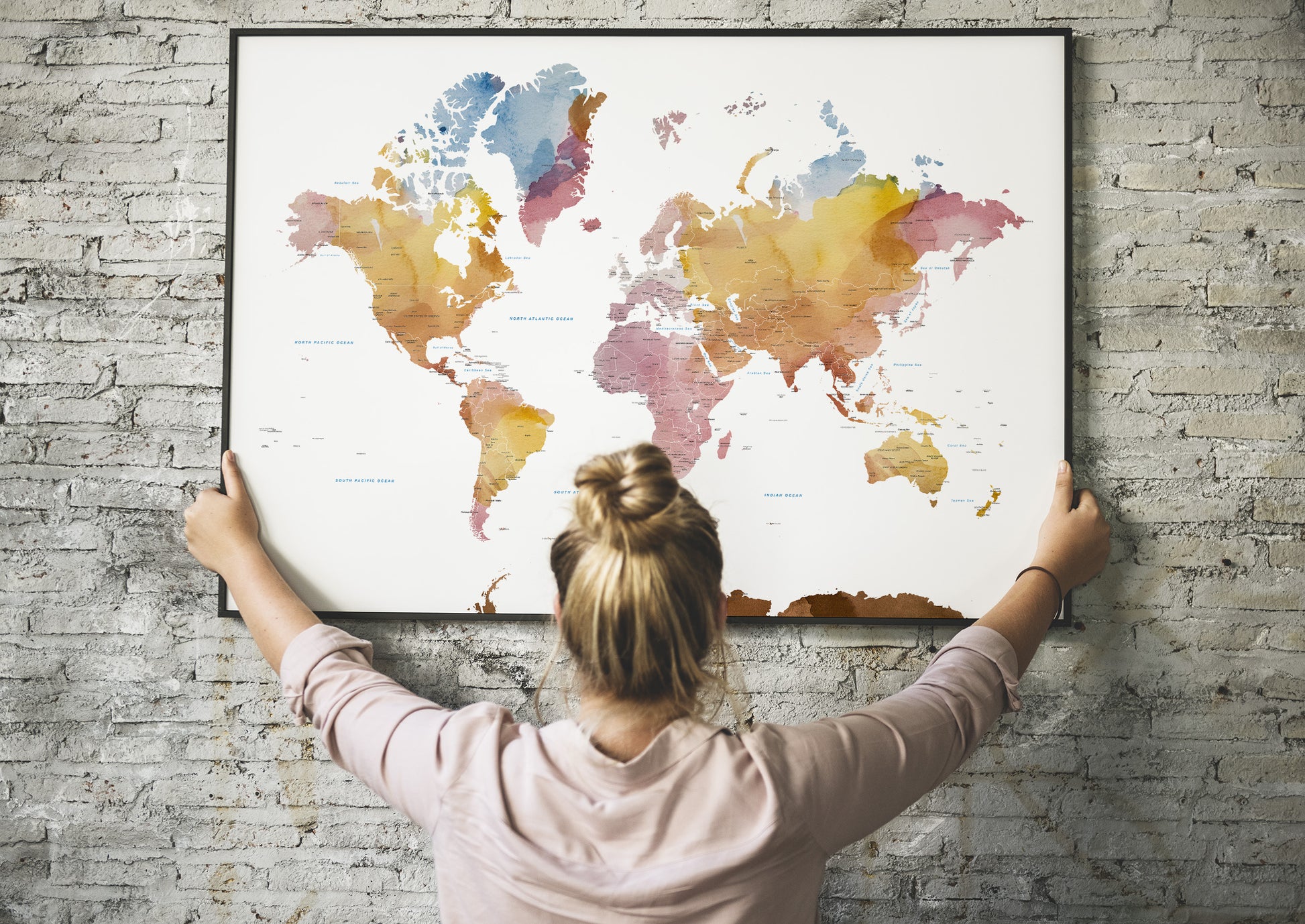 A woman with her hair in a bun hangs a large framed world map poster on a gray brick wall, viewed from behind as she holds the frame against the wall.