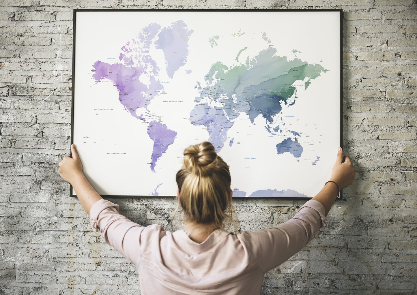 A woman with her hair in a bun hangs a large framed world map poster on a gray brick wall, viewed from behind her.