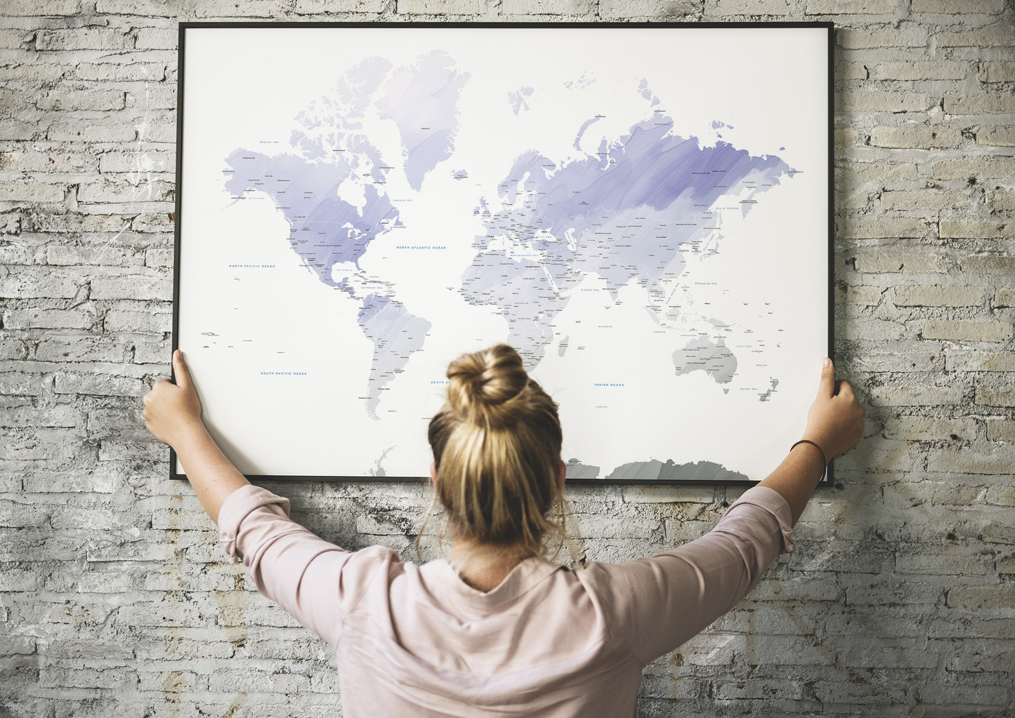 A woman seen from behind holding a large framed world map poster against a light gray brick wall.