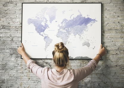 A woman seen from behind holding a large framed world map poster against a light gray brick wall.