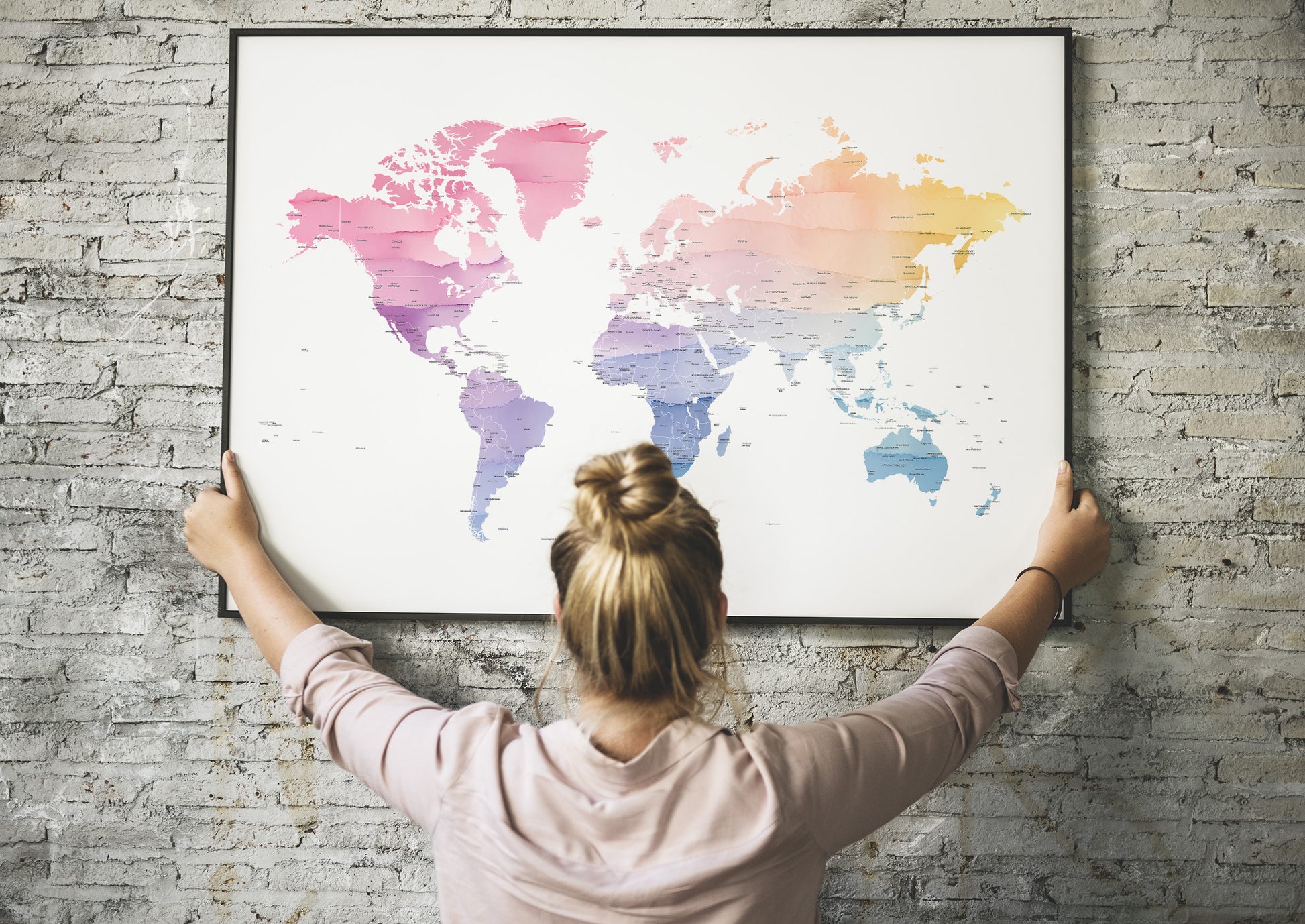 A woman with her hair in a bun hangs a large framed world map poster on a gray brick wall, viewed from behind her.