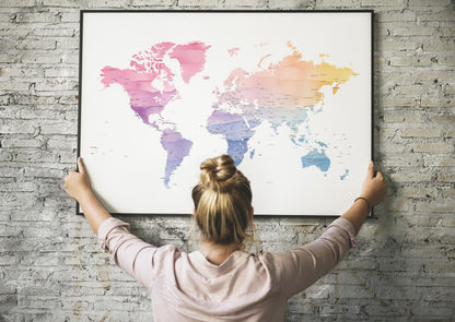 A woman with her hair in a bun hangs a large framed world map poster on a gray brick wall, viewed from behind her.