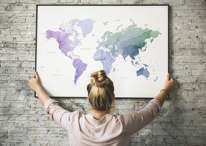 A woman with her hair in a bun hangs a large framed world map poster on a light gray brick wall, viewed from behind her.