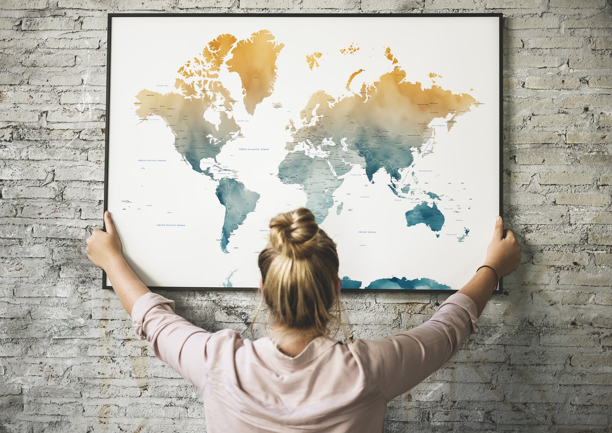 A woman with her hair in a bun is seen from behind, holding a large framed world map poster against a gray brick wall. The map features a watercolor design with gold, orange, and teal landmasses on a white background.