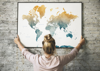 A woman with her hair in a bun is seen from behind, holding a large framed world map poster against a gray brick wall. The map features a watercolor design with gold, orange, and teal landmasses on a white background.