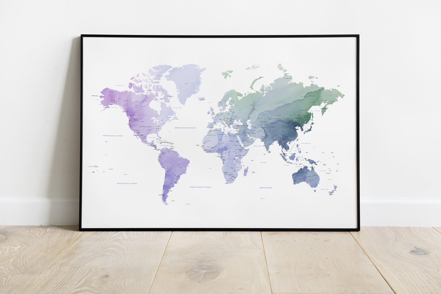 A framed world map art print with a watercolor design in shades of purple, blue, and green on a white background, leaning against a white wall on a light wood floor.