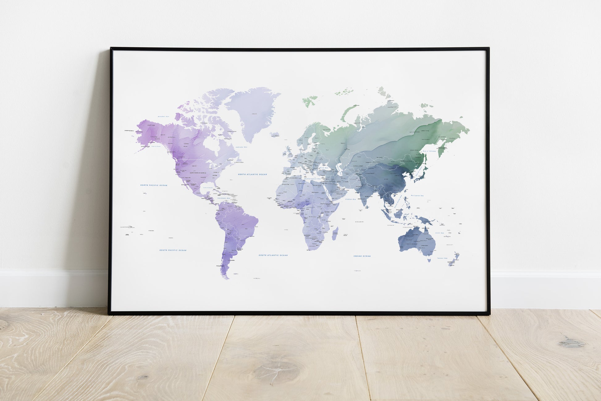 A framed world map art print with a watercolor design in shades of purple, blue, and green on a white background, leaning against a white wall on a light wood floor.