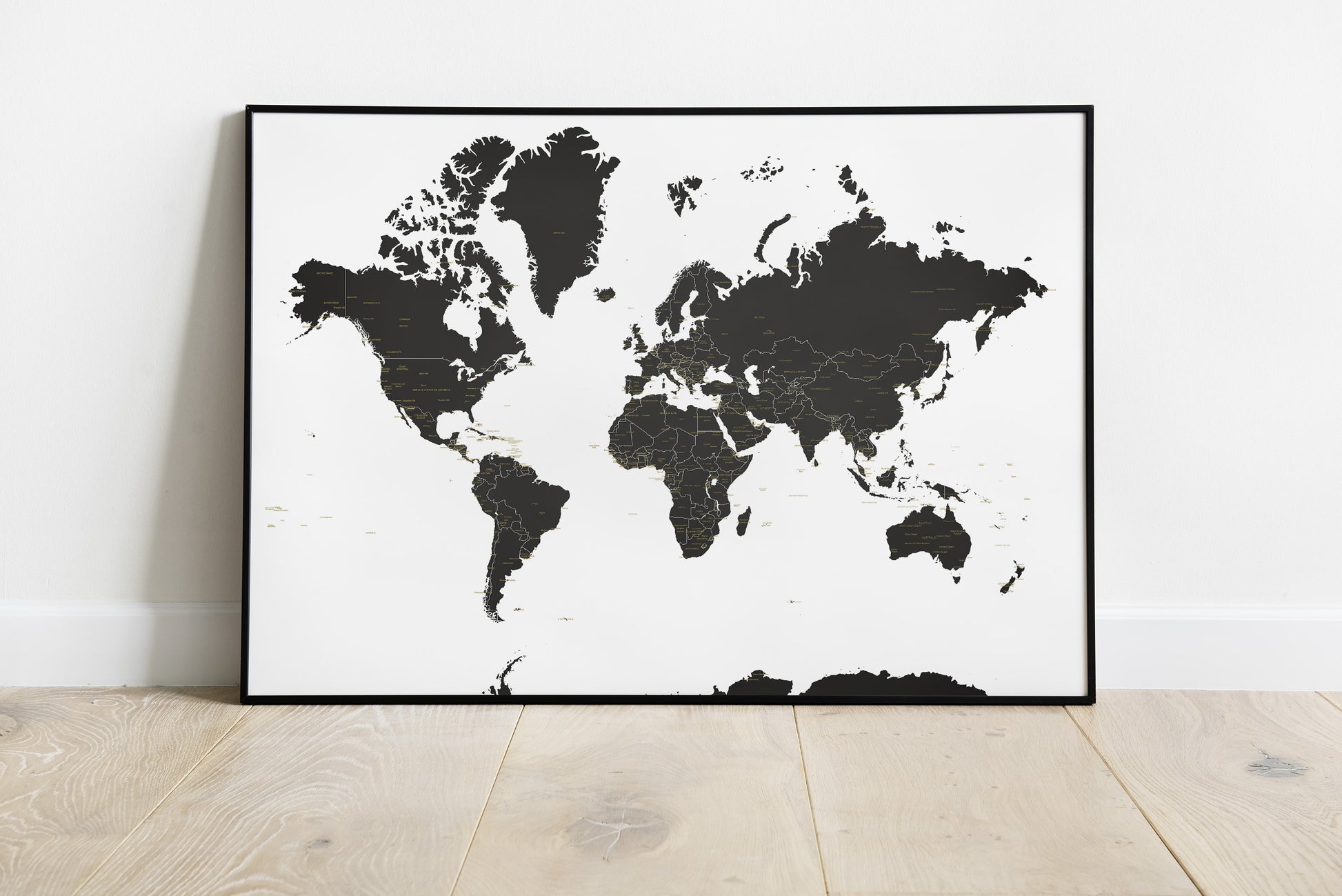 A framed world map poster with black landmasses on a white background, leaning against a white wall on a light wood floor.