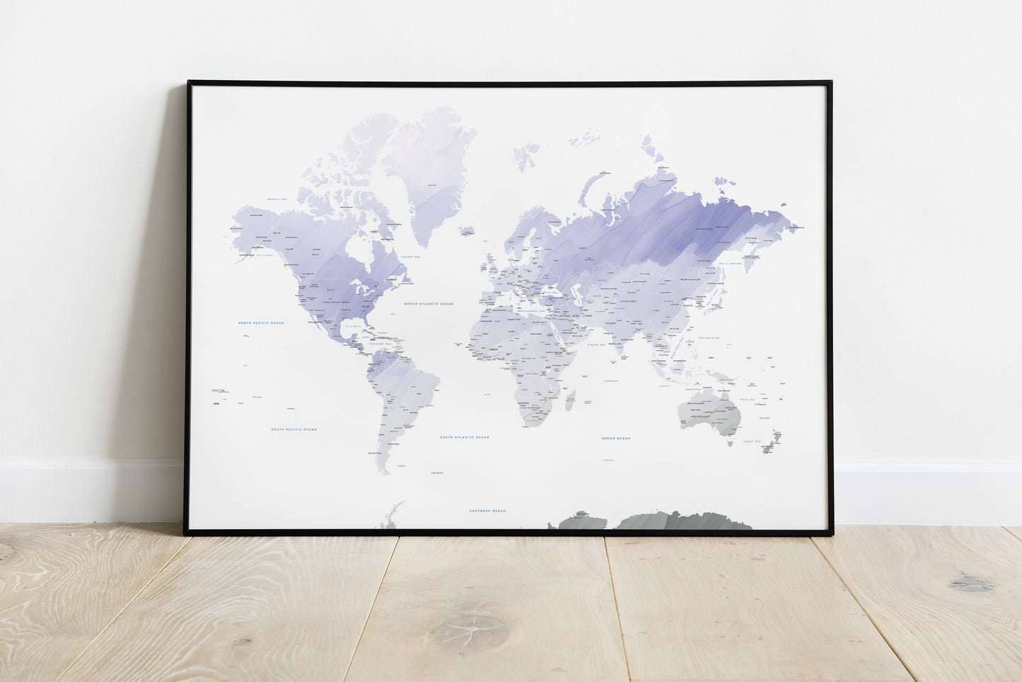 A framed world map art print with watercolor-style continents in shades of lavender, purple, and gray on a white background, leaning against a white wall on a light wood floor.