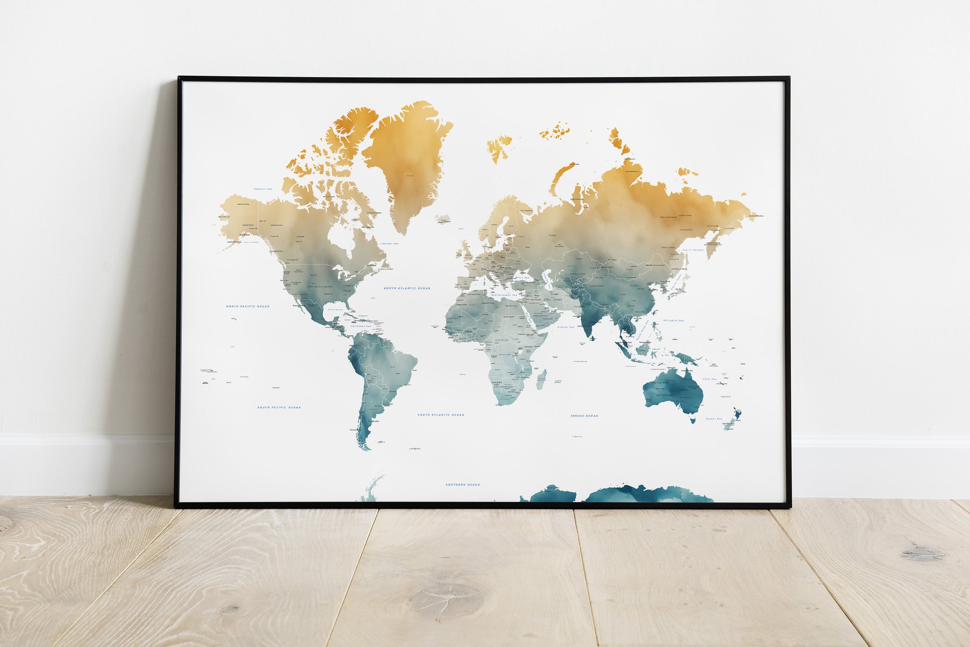 A framed world map watercolor print with gold, teal, and blue landmasses on a white background, leaning against a white wall on a light wood floor.