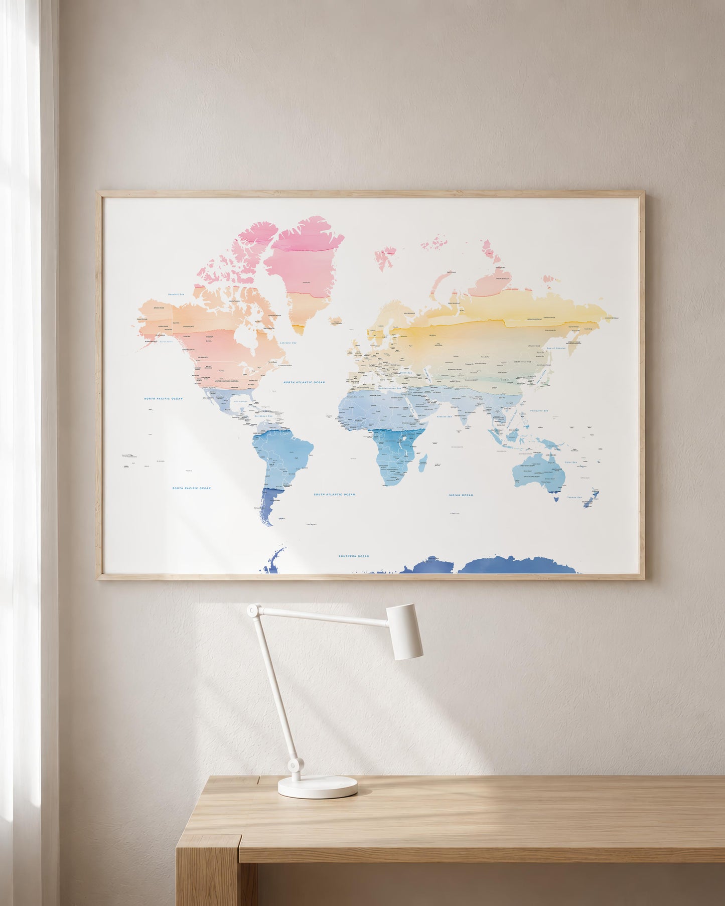 A framed world map poster with pastel watercolor-style continents hangs on a beige wall above a light wood desk with a white desk lamp.