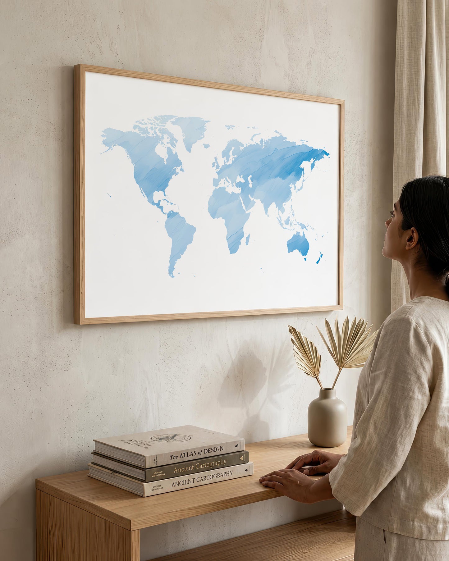 A woman in a beige linen outfit stands beside a light wood console table, looking at a large framed world map print with blue watercolor continents on a white background, hanging on a textured beige wall.