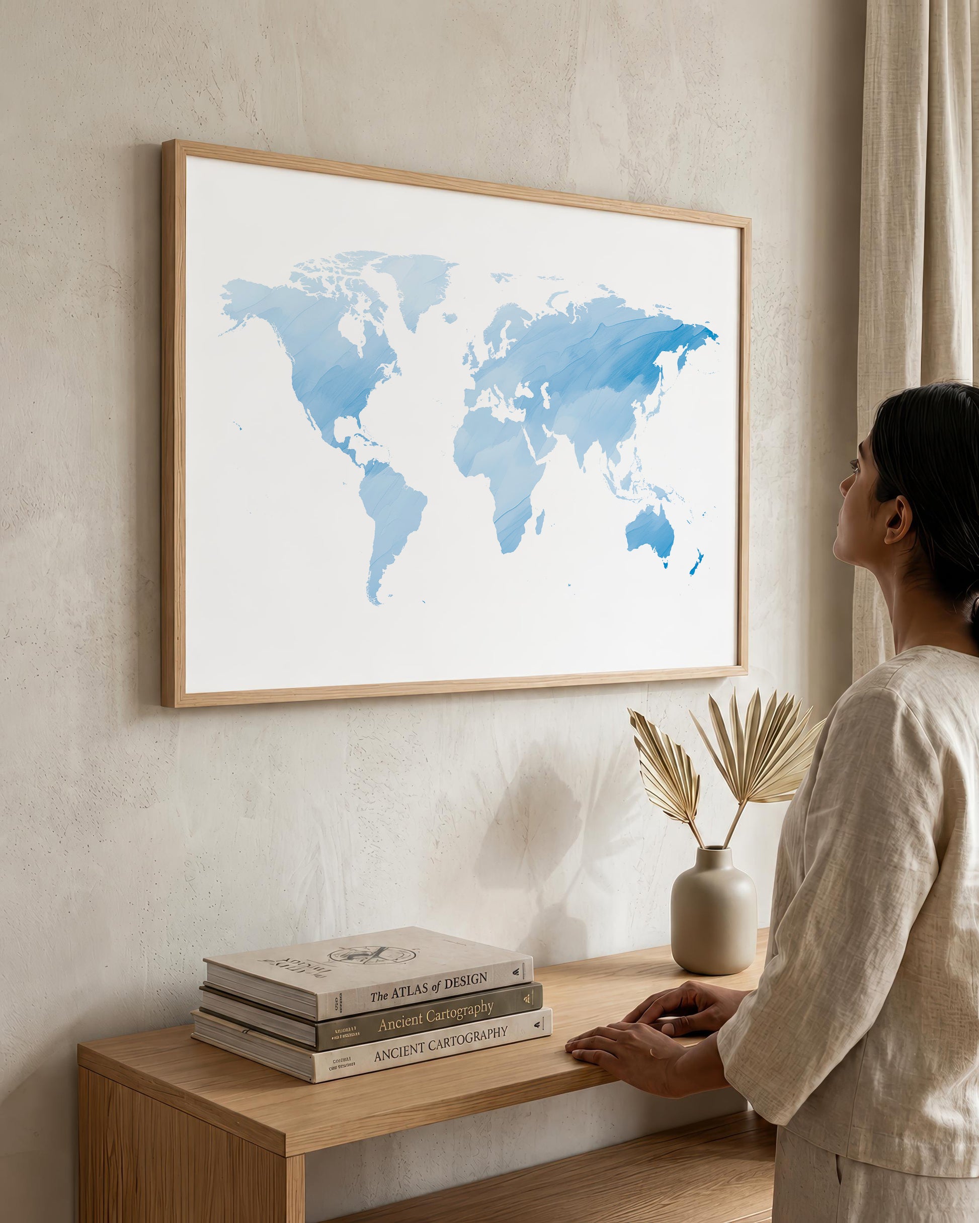 A woman in a beige linen outfit stands beside a light wood console table, looking at a large framed world map print with blue watercolor continents on a white background, hanging on a textured beige wall.