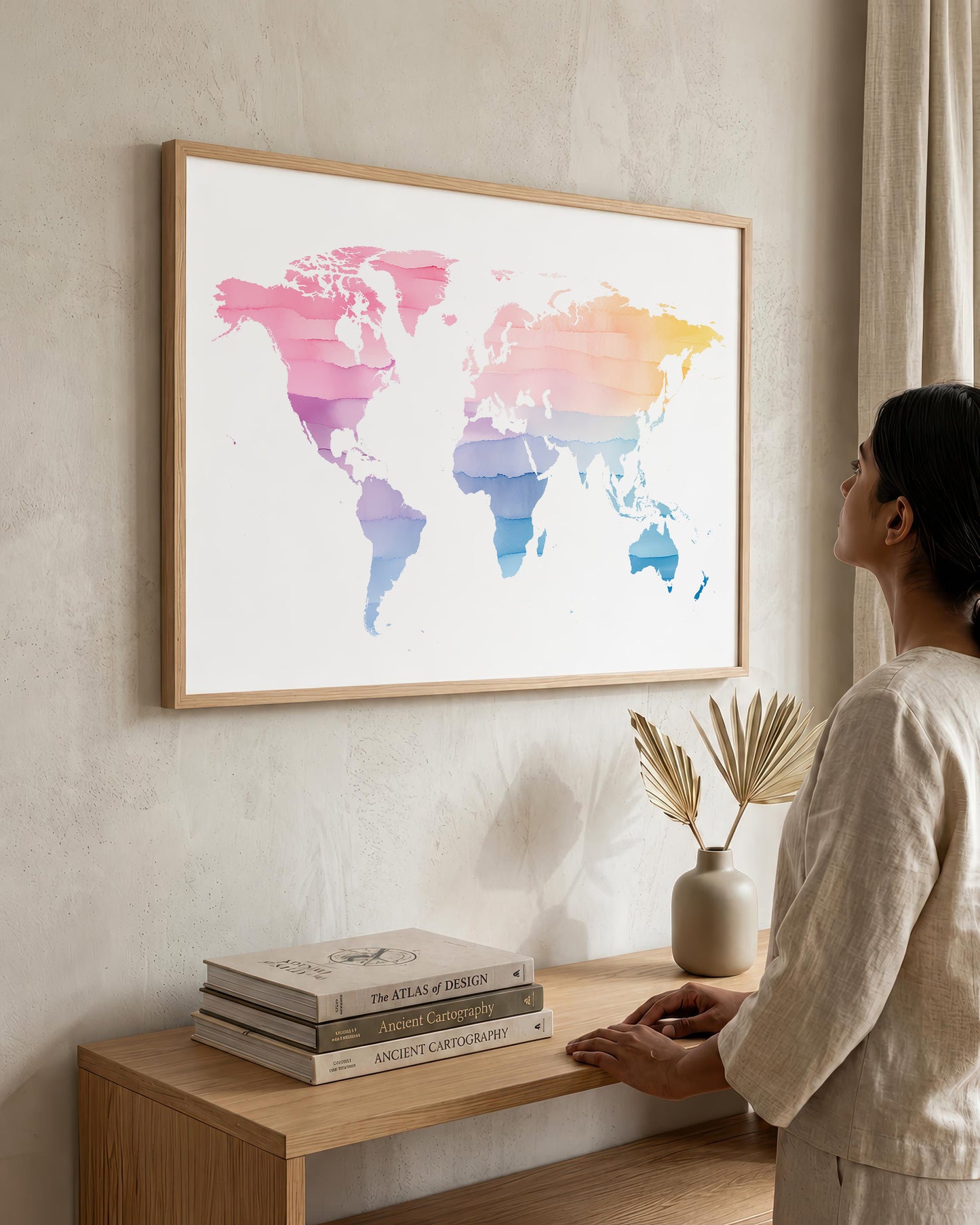 A woman in a beige linen outfit stands beside a wooden console table, looking at a large framed world map art print hanging on a textured beige wall. The map features a watercolor design with pink, purple, blue, and yellow tones on a white background, enclosed in a light wood frame.
