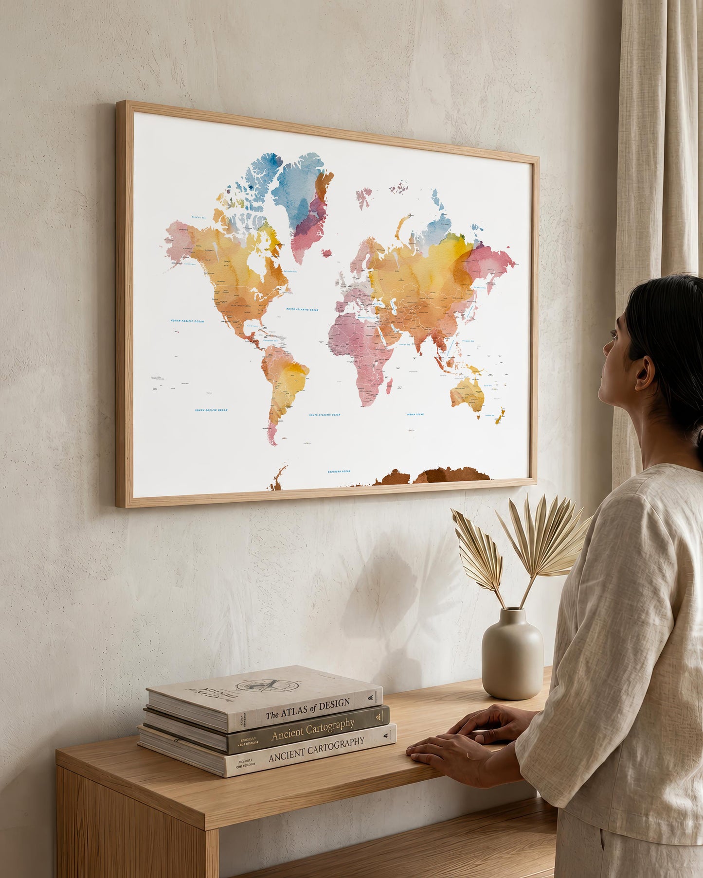 A woman in a neutral-toned outfit stands beside a wooden console table, looking at a large framed world map art print hanging on a textured beige wall. The map features watercolor continents in soft blue, pink, orange, and yellow tones on a white background, enclosed in a light wood frame.