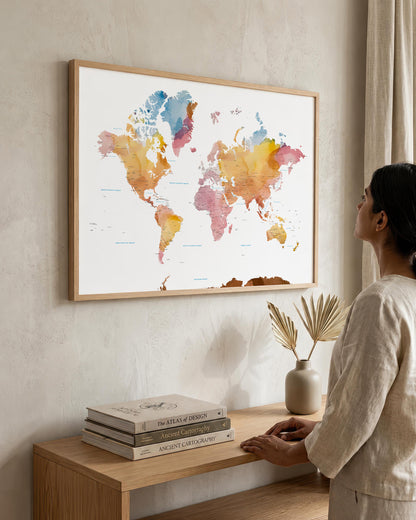 A woman in a neutral-toned outfit stands beside a wooden console table, looking at a large framed world map art print hanging on a textured beige wall. The map features watercolor continents in soft blue, pink, orange, and yellow tones on a white background, enclosed in a light wood frame.