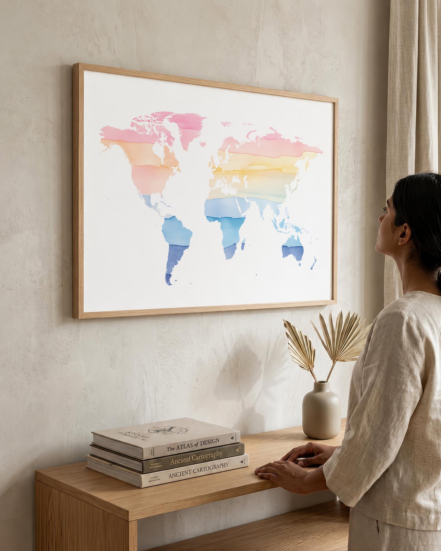 A woman in a neutral-toned outfit stands beside a wooden console table, looking at a large framed world map art print hanging on a textured beige wall. The map features watercolor-style continents in pink, orange, yellow, and blue on a white background, enclosed in a light wood frame.