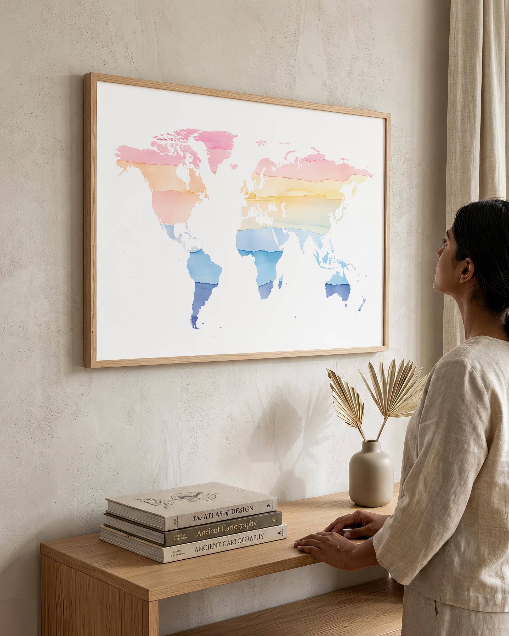 A woman in a neutral-toned outfit stands beside a wooden console table, looking at a large framed world map art print hanging on a textured beige wall. The map features watercolor-style continents in pink, orange, yellow, and blue on a white background, enclosed in a light wood frame.