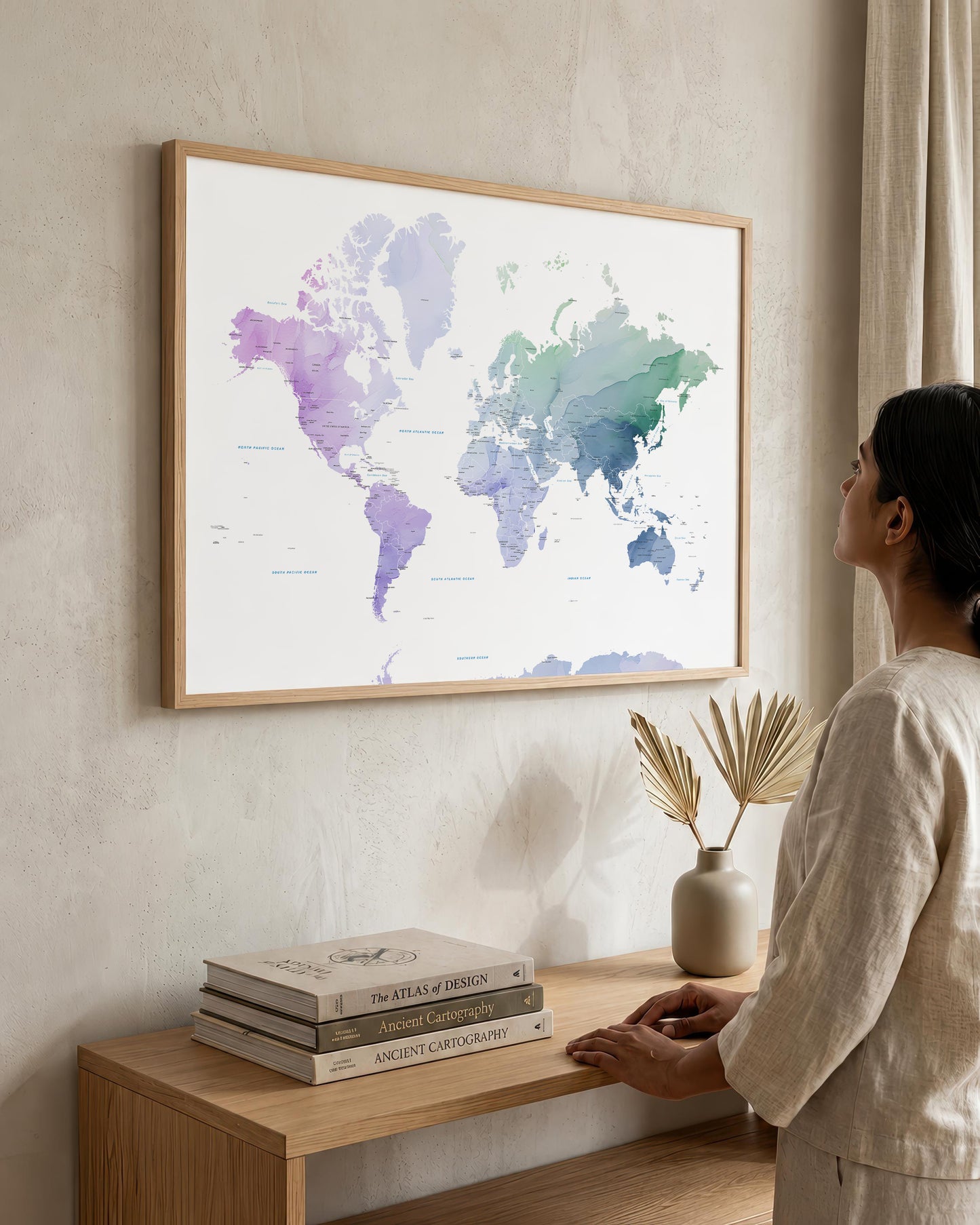 A woman in a neutral-toned outfit stands beside a wooden console table, looking at a large framed world map hanging on a textured beige wall. The map features watercolor-style continents in soft purple, green, and blue tones on a white background, enclosed in a light wood frame.