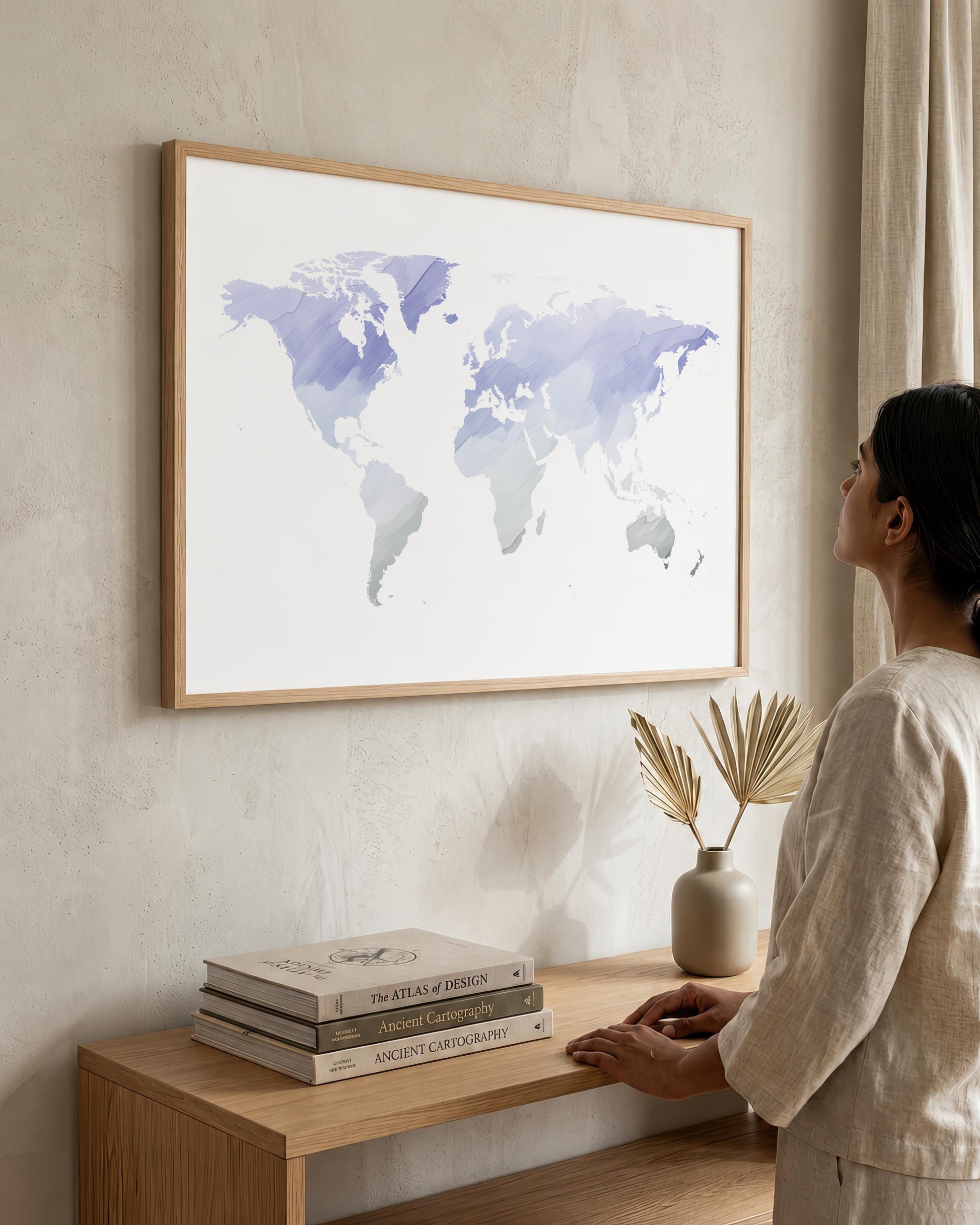A woman in a beige linen outfit stands beside a wooden console table, looking at a large framed world map print hanging on a textured beige wall. The print features watercolor continents in shades of blue, purple, and gray on a white background, enclosed in a light wood frame.