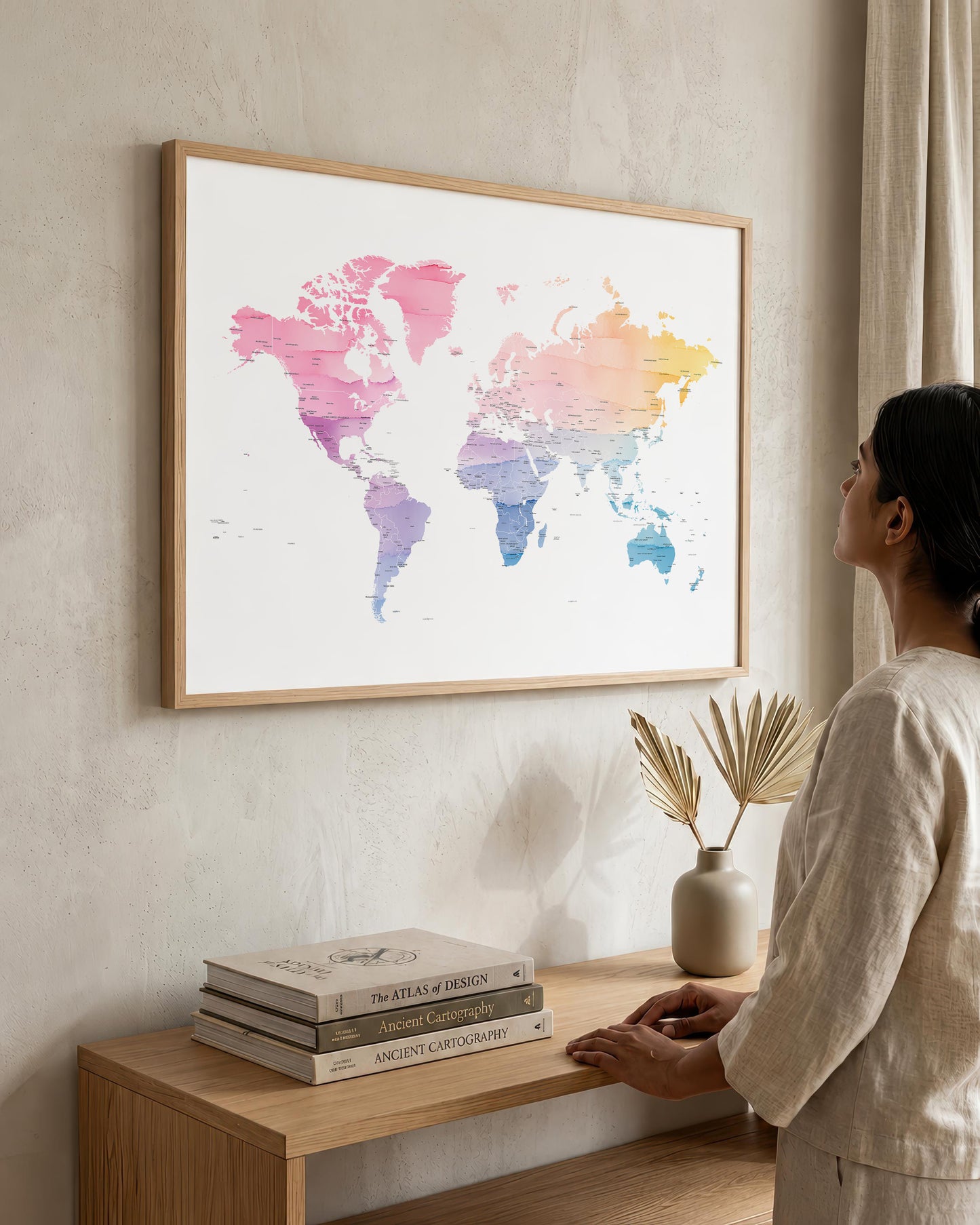A woman in a neutral-toned outfit stands beside a wooden console table, looking at a large framed world map art print hanging on a textured beige wall. The map features a watercolor design with pink, purple, blue, and yellow hues on a white background, enclosed in a light wood frame.