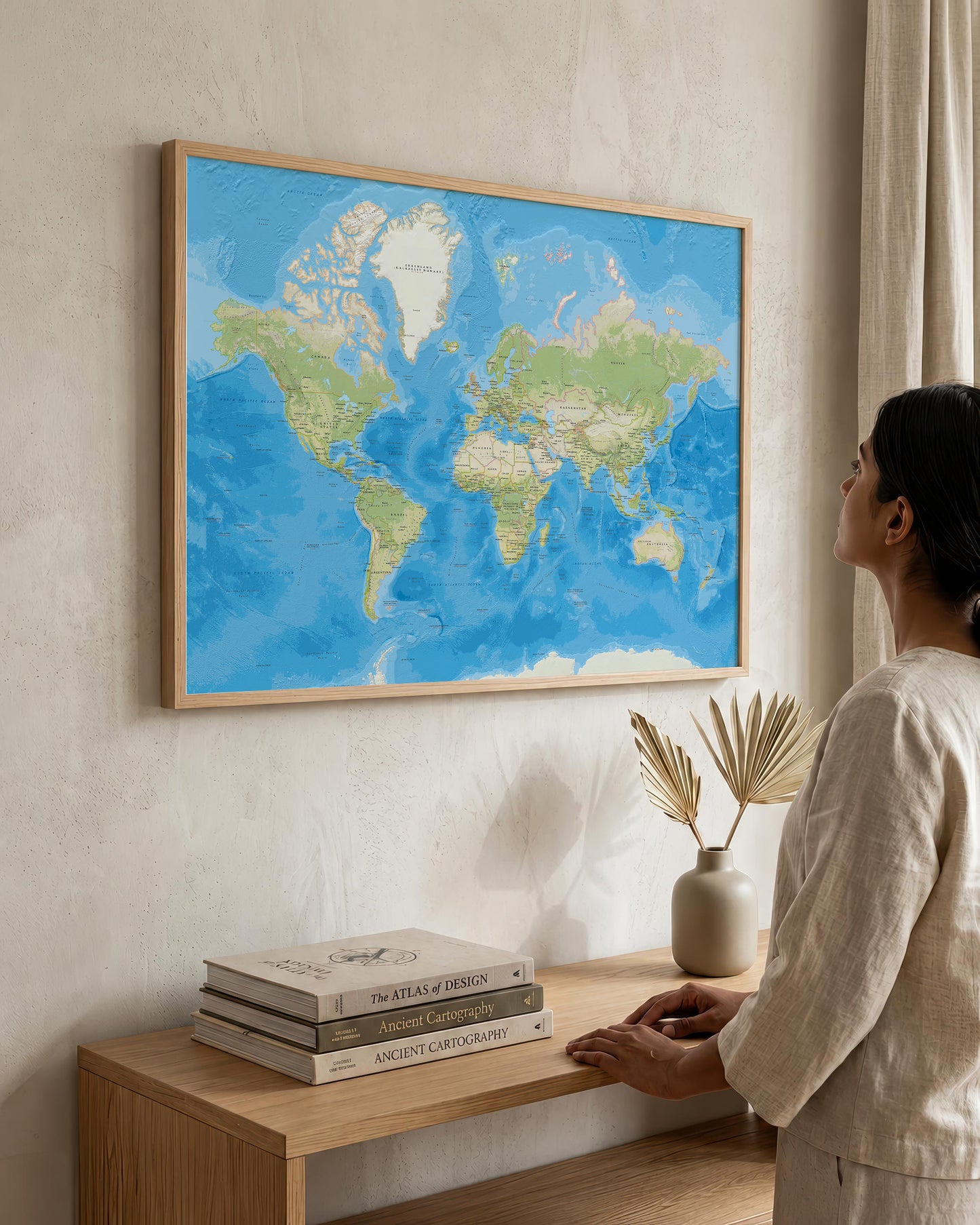 A woman in a beige outfit stands beside a wooden console table, looking at a large framed world map hanging on a textured beige wall. The map features blue oceans and green landmasses, enclosed in a light wood frame.