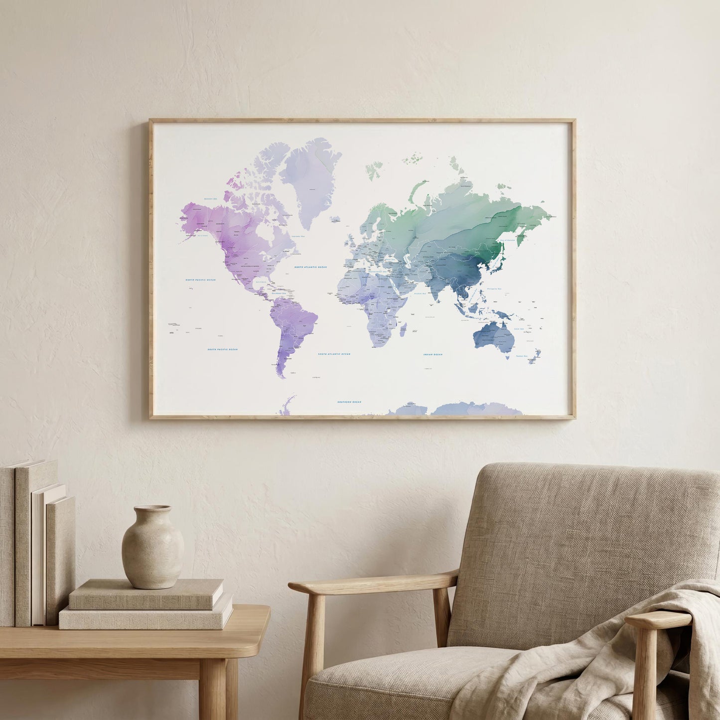 A framed world map poster with a watercolor design in shades of purple, blue, and green, hanging on a light beige wall above a modern armchair and a small side table with books and a vase.