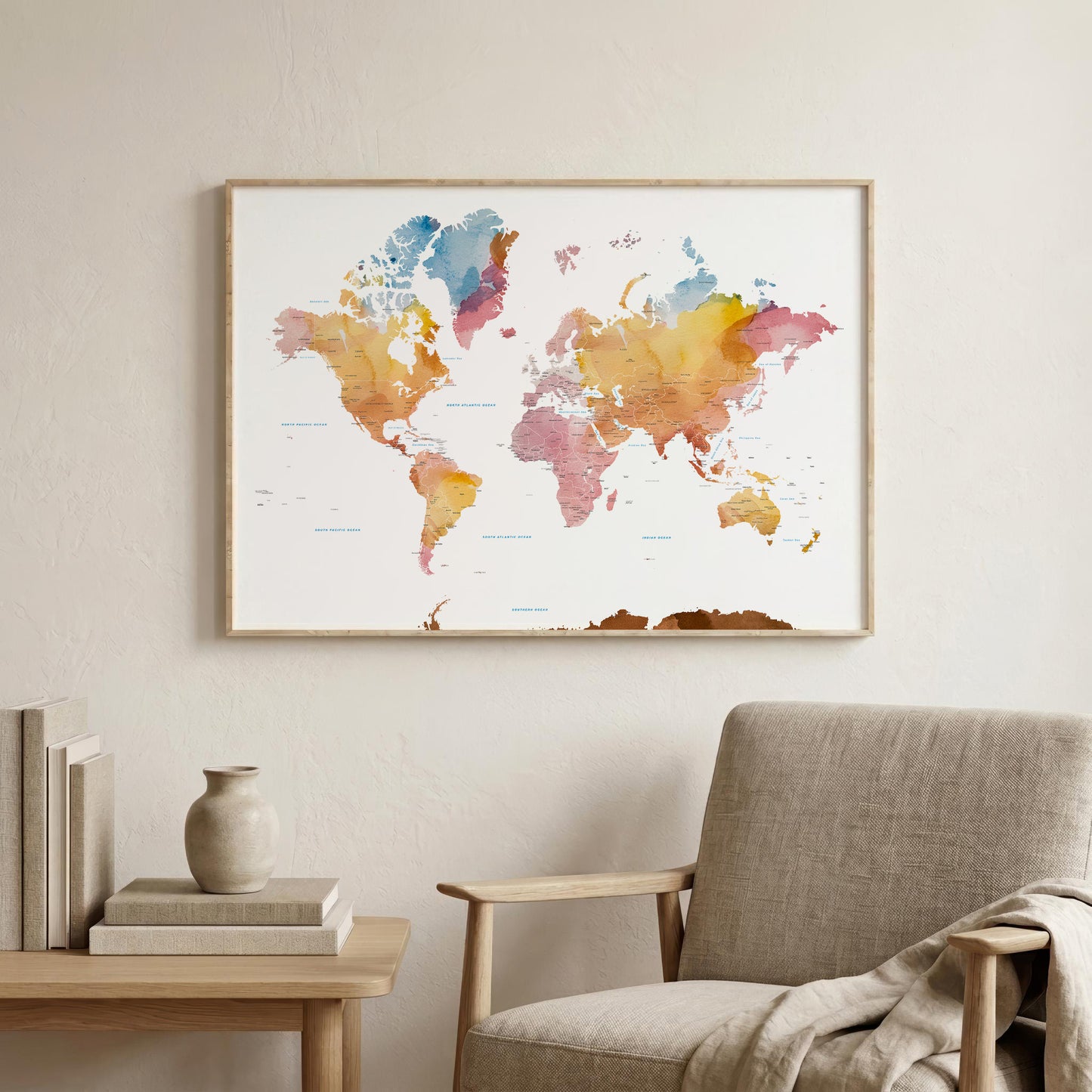 A framed world map watercolor print hangs on a light beige wall above a wooden side table and a gray upholstered armchair. The map features soft watercolor continents in orange, yellow, pink, blue, and purple tones on a white background, enclosed in a thin natural wood frame.