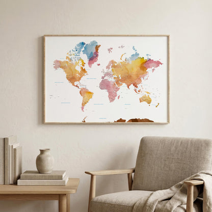 A framed world map watercolor print hangs on a light beige wall above a wooden side table and a gray upholstered armchair. The map features soft watercolor continents in orange, yellow, pink, blue, and purple tones on a white background, enclosed in a thin natural wood frame.