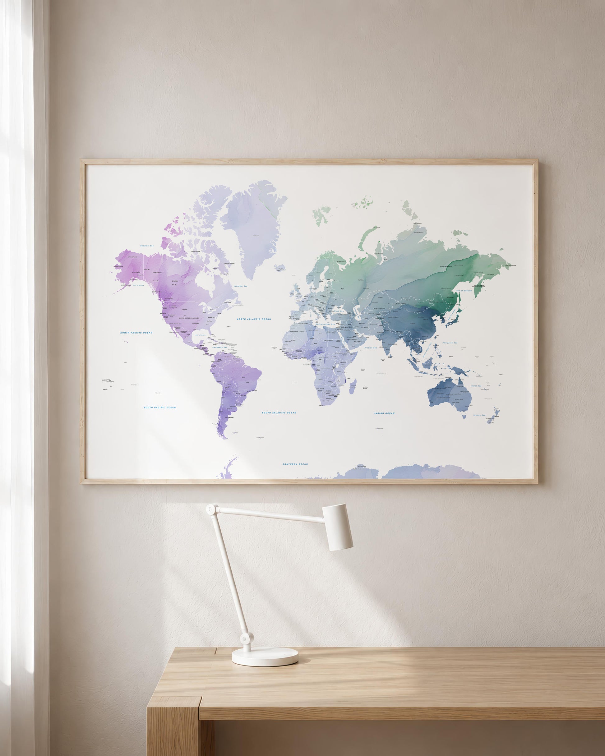 A framed world map poster with a watercolor design in shades of purple, blue, and green, hanging on a beige wall above a wooden desk with a white desk lamp.
