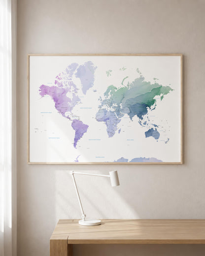 A framed world map poster with a watercolor design in shades of purple, blue, and green, hanging on a beige wall above a wooden desk with a white desk lamp.
