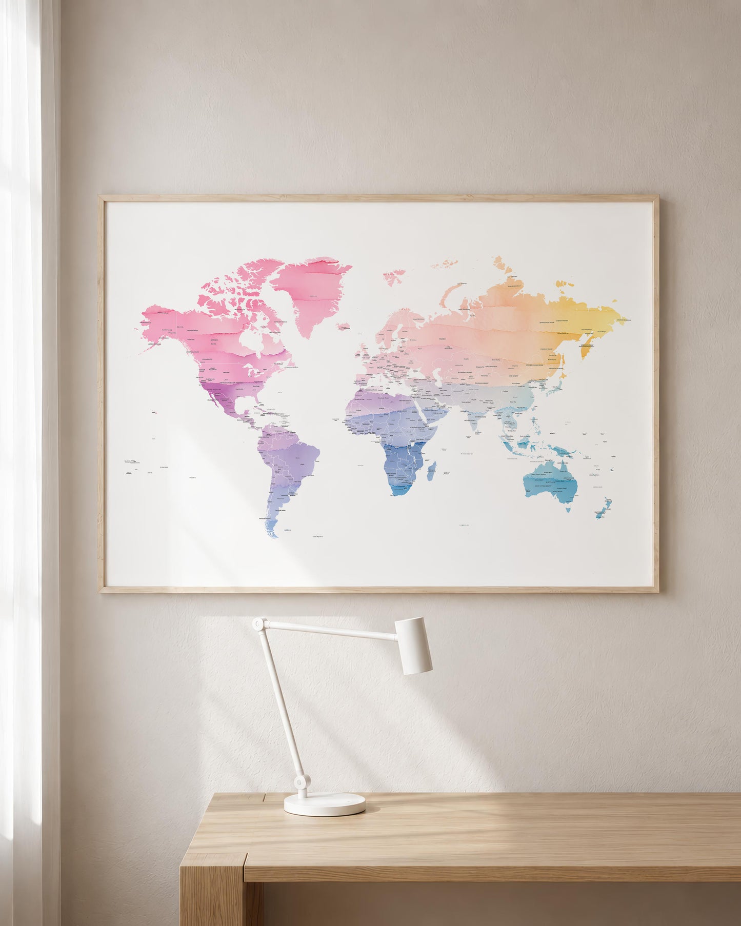 A framed world map art print with a watercolor gradient in pink, purple, blue, and yellow tones, hanging on a light beige wall above a wooden desk with a white desk lamp.