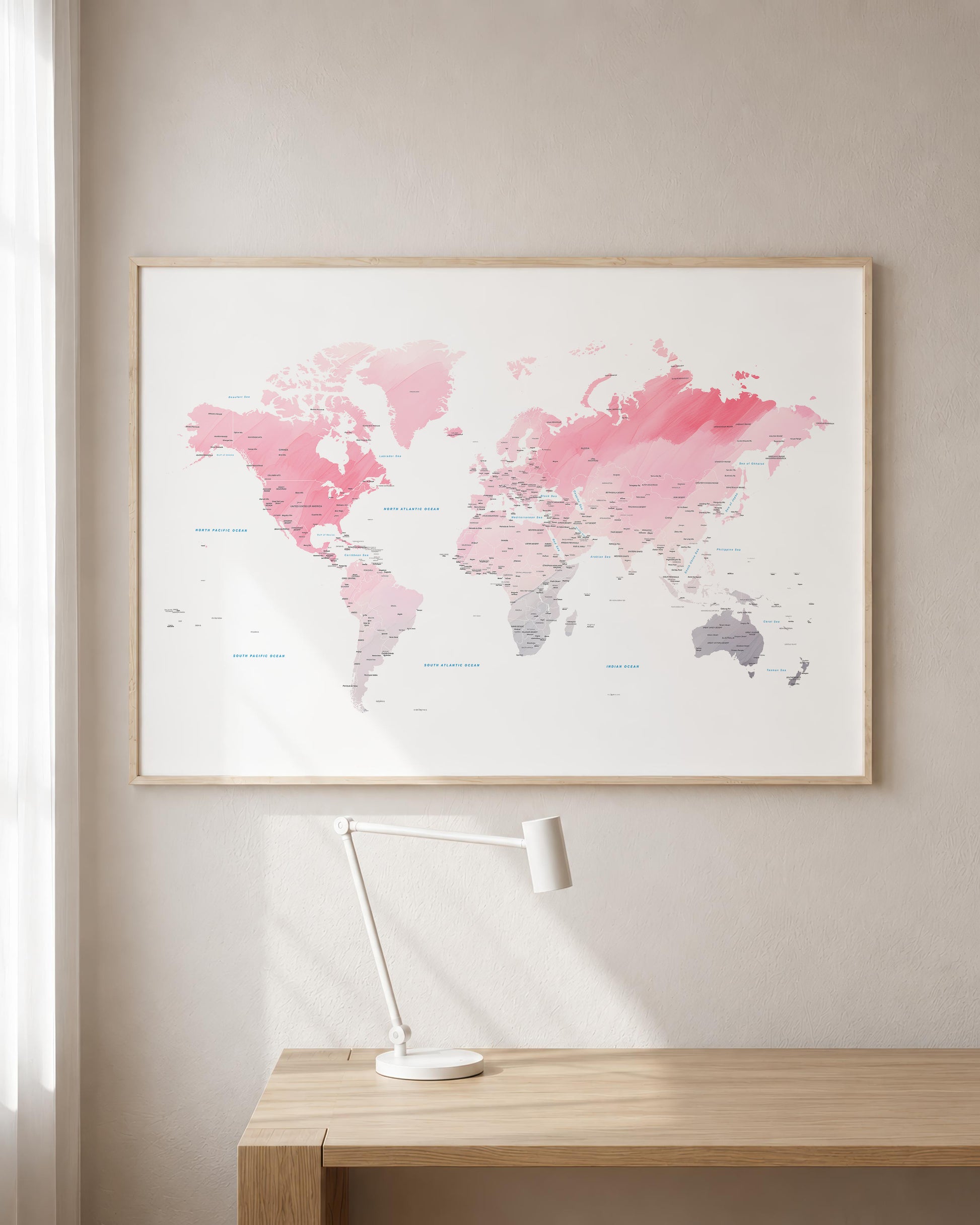 A framed world map art print with pink and gray watercolor continents on a white background, hanging on a beige wall above a light wood desk with a white desk lamp.