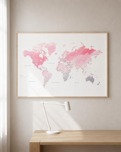 A framed world map art print with pink and gray watercolor continents on a white background, hanging on a beige wall above a light wood desk with a white desk lamp.