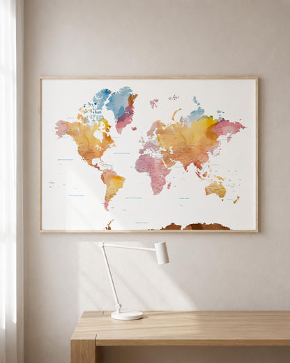 A framed world map watercolor print hangs on a light beige wall above a wooden desk with a white desk lamp.