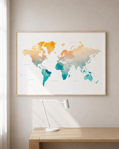 A framed world map poster with a watercolor design in orange, beige, and teal tones, hanging on a light gray wall above a wooden desk with a white desk lamp.