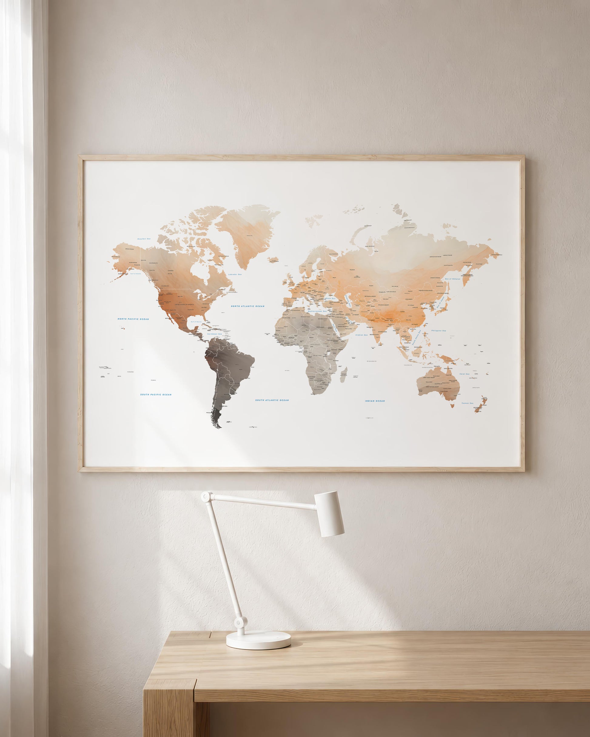 A framed world map wall art print hangs on a beige wall above a light wood desk with a white desk lamp. The map features watercolor-style continents in muted peach, tan, gray, and brown tones on a white background, enclosed in a thin natural wood frame.