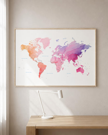 A framed world map poster with a watercolor design in pink, purple, orange, and blue tones, displayed on a beige wall above a wooden desk with a white lamp.