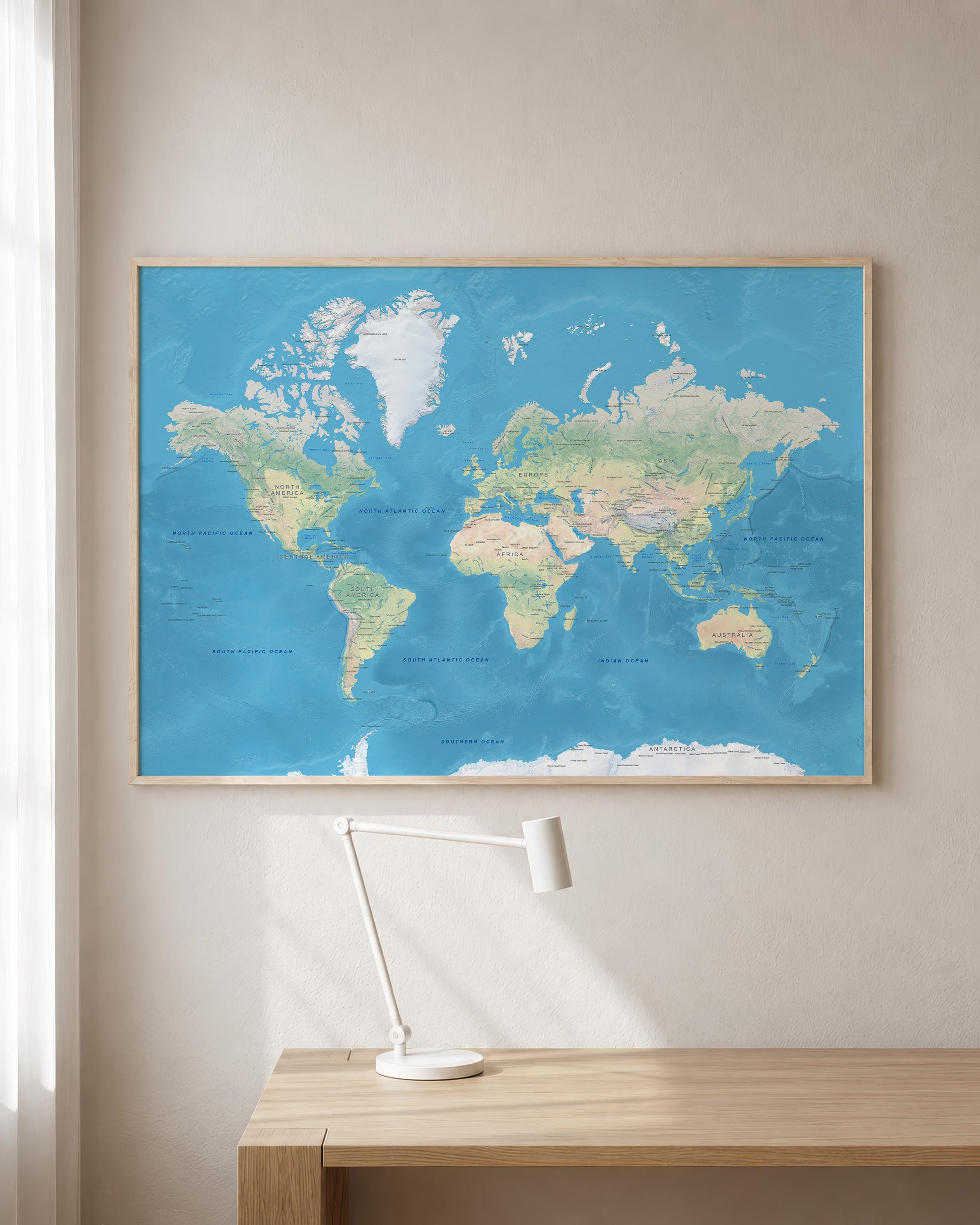 A framed world map poster with a light blue ocean and beige landmasses, hanging on a beige wall above a light wood desk with a white desk lamp.