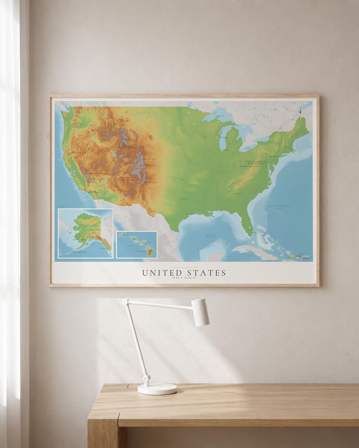 A framed United States topographic map print hanging on a beige wall above a light wood desk with a white desk lamp.