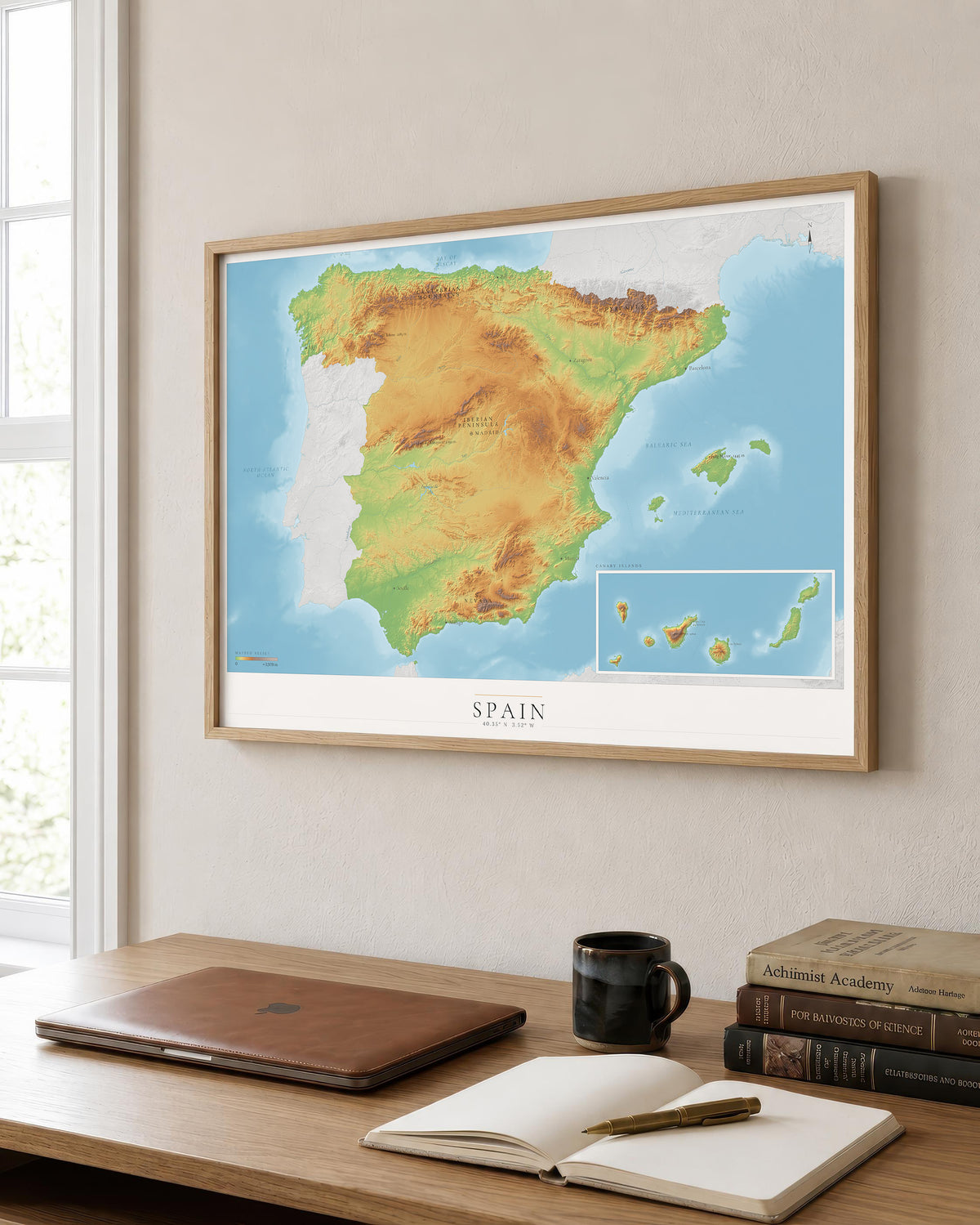 A framed topographic map of Spain and Portugal hanging on a light beige wall above a wooden desk with a laptop, notebook, and books.
