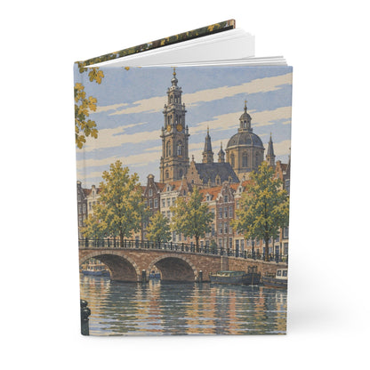 A hardcover notebook or journal standing upright, featuring a watercolor-style cityscape print on the cover with a canal, bridge, trees, and historic buildings under a blue sky.