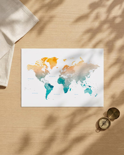 A watercolor world map art print with orange, teal, and peach tones on a white background, styled in a flat lay on a light wood surface with leaf shadows, a folded cream cloth, and a small brass compass.